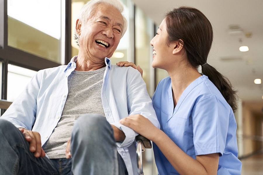 Home Health Care Insurance - Smiling Caregiver Taking Care Of Elderly Man At Nursing Home